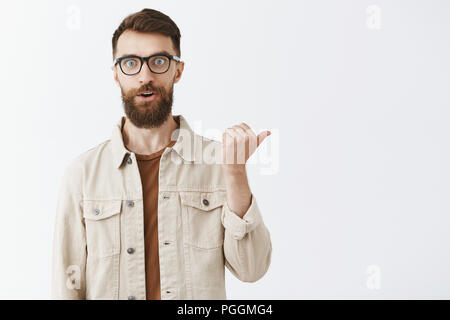 Studio shot of impressed and thrilled charming adult male customer asking question about awesome product in shop staring at camera excited pointing right with thumb holding breath from excitement Stock Photo