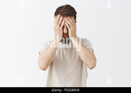 Studio shot of man suffering from painful feelings griefing bending head down covering face with palms being fed up and depressed, standing tired after working all night over gray background Stock Photo