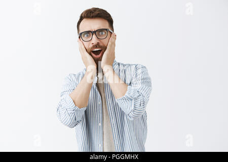 Waist-up shot of impressed and surprised sociable good-looking male with long beard and moustache holding palms on cheeks and staring at camera hearing shocking rumor over gray background Stock Photo