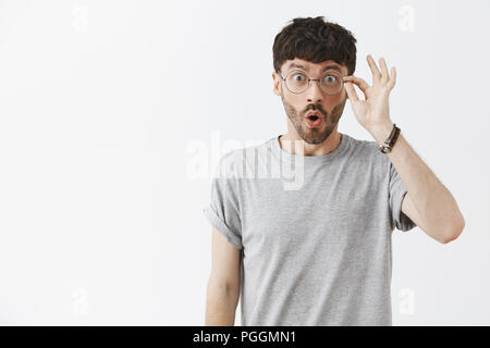 Waist-up shot of enthusiastic impressed dark-haired guy with beard in stylish transparent glasses touching rim of eyewear and folding lips from joy and excitement staring at amazing advertisement Stock Photo