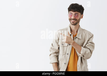 Waist-up shot of enthusiastic creative stylish male in pink sunglasses and trendy outfit pointing and gazing at upper left corner pleased and joyful enjoying spending great time in artistic company Stock Photo
