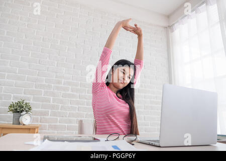 Asian woman stretching arm up when working on laptop at white brick wall in home.Work at home concept.relax after hard work Stock Photo