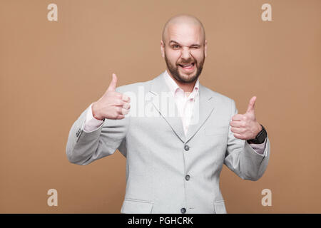 Portrait of funny satisfied handsome middle aged bald bearded businessman in classic light gray suit with thumbs up standing, looking at camera. indoo Stock Photo