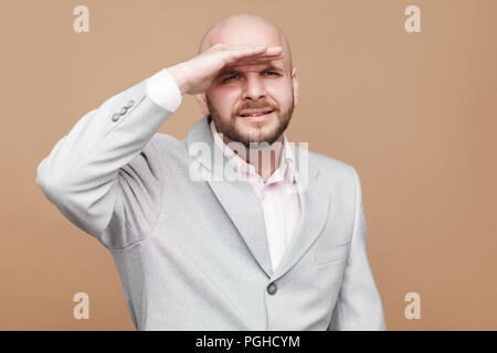 Portrait of confused handsome middle aged bald bearded businessman in classic light gray suit standing and looking too far with gesture hands on his h Stock Photo