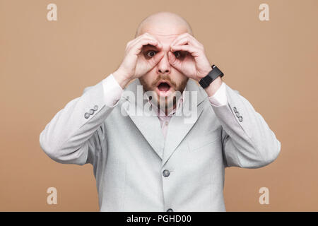Wondered middle aged bald bearded businessman in classic light gray suit standing with binoculars gesture and looking at camera with shocked face. ind Stock Photo