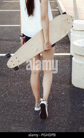 Rear view of skater girl walking away holding her longboard behind her back Stock Photo