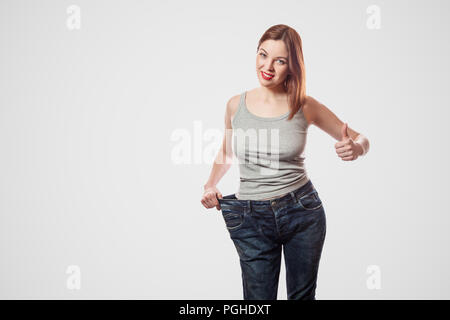 portrait of happy beautiful slim waist of young woman in big jeans and gray top showing successful weight loss, indoor, studio shot, isolated on light Stock Photo