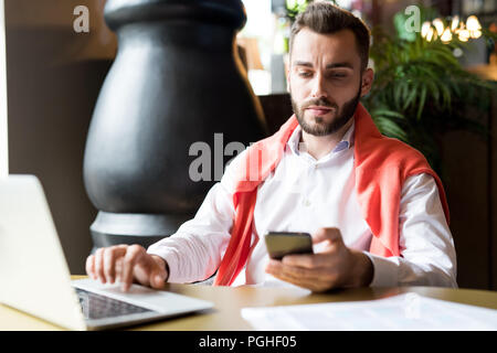 Portrait of handsome bearded businessman holding smartphone typing text messages while working at table in cafe Stock Photo