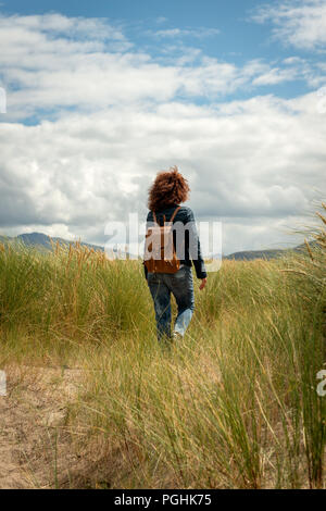 Young beautiful woman with red curly hair wearing casual  blue jacket and blue jeans walking on coastal sand dune through marram grass on a sunny day. Stock Photo