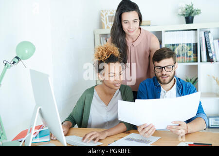 Multi-ethnic group of contemporary young business people discussing documents while working at desk in modern office, all dressed in business casual Stock Photo