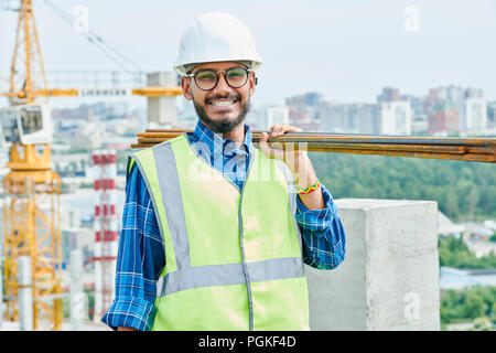 Waist up portrait of cheerful Middle-Eastern construction worker wearing harhat and holding metal bars smiling at camera while enjoying work on site Stock Photo