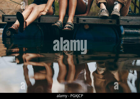 Friends on a holiday sitting together on a floating dock made out of plastic barrels. Friends sitting near lake with their reflection in the water. Stock Photo