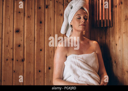 Woman with towel wrapped around her body and head sitting in sauna spa peacefully. Woman relaxing in a wooden spa with eyes closed. Stock Photo