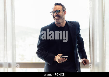Smiling portrait of mature businessman standing by a window with mobile phone. Middle aged man in business suit looking away and smiling. Stock Photo