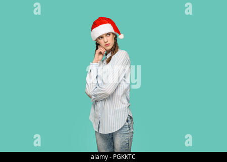 New year concept. thoughtful beautiful girl in striped light blue shirt in red christmas cap standing crossed hands, confused face and thinking. indoo Stock Photo