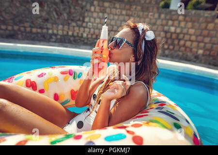 Beautiful young woman in swimming pool swims on inflatable ring donut and has fun with bottle of cocktail on vacation. Stock Photo
