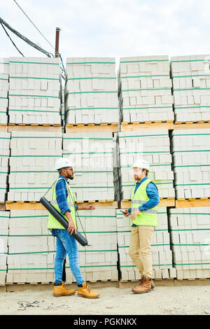 Full length portrait of two construction workers discussing project standing against concrete blocks on site Stock Photo
