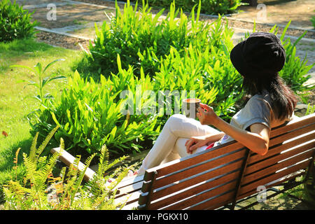Young woman relaxing on wooden bench in vibrant green garden holding a cup of hot coffee in the morning Stock Photo