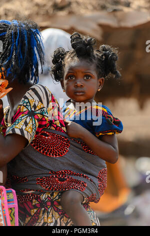 BOHICON, BENIN - JAN 12, 2017: Unidentified Beninese little curly  girl is carried by her mother at the local market. Benin children suffer of poverty Stock Photo