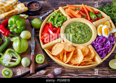 green salsa verde and mexican set of chopped fresh vegetables, hard boiled eggs and chips in bowls on old rustic wooden board with ingredients, view f Stock Photo