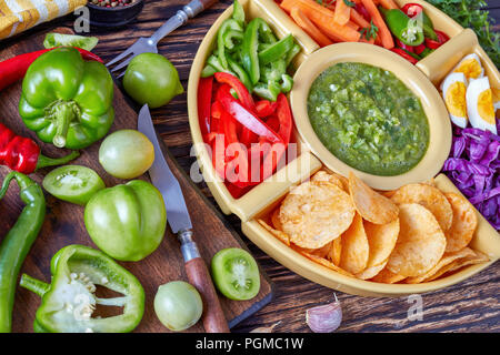 green salsa verde and mexican set of chopped fresh vegetables, hard boiled eggs and chips in bowls on old rustic wooden table with ingredients, horizo Stock Photo