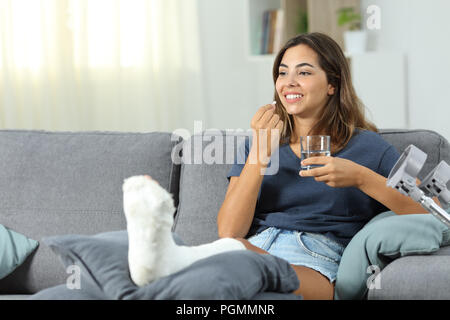 Happy disabled woman taking a painkiller pill sitting on a couch in the living room at home Stock Photo