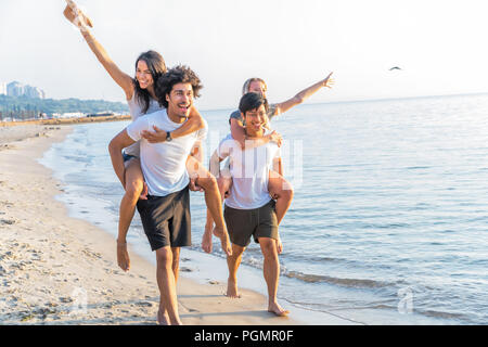 Group of friends walking along the beach, with men giving piggyback ride to girlfriends. Happy young friends enjoying a day at beach