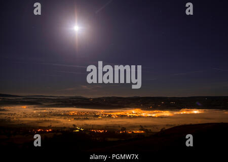 Streetlights of Ruthin at night glowing through fog in the Vale of Clwyd, North Wales, with the moon above Stock Photo