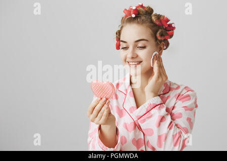 Glamorous attractive woman with hair-curlers on head, wearing pyjamas and holding heart-shaped mirror, smiling while being glad she has good skin, taking off makeup with cotton pad over gray wall Stock Photo