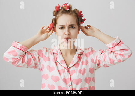 Bothered and focused cute caucasian girl in hair-curlers, wearing pyjamas while in bathroom, frowning and showing tongue, being concentrated on taking off her wavers, standing over gray background. Stock Photo