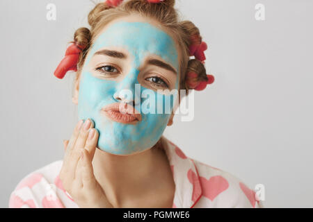 Close-up portrait of funny white woman with hair-curlers, wearing nightwear and folding lips while applying face mask with hand, looking at camera as if in mirror. Girl decided to take day off. Stock Photo