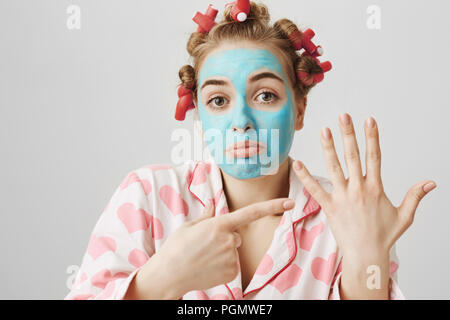 Portrait of upset funny housewife in hair-curlers pyjamas and blue face mask pointing at left arm, asking where is her ring, standing over gray background. Girl when boyfriend will make her proposal Stock Photo