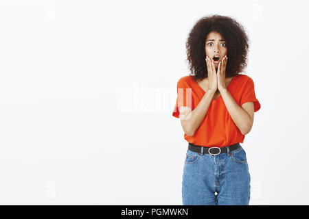 Indoor shot of worried shocked attractive dark-skinned woman with afro hairstyle, dropping jaw from amazement and surprise, gasping, holding hands on cheeks, standing against gray background Stock Photo