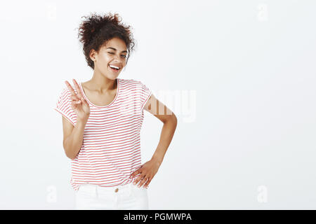 Indoor shot of confident happy and outgoing woman with tanned skin, holding hand on hip, showing victory or peace gesture and winking with broad smile at camera, posing over gray background Stock Photo