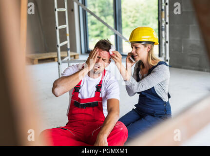 Accident of a male worker at the construction site. A woman with smartphone helping her injured colleague, wiping his forehead. Stock Photo
