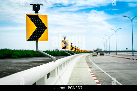 Black and yellow arrow on curve traffic sign on the bridge with solar cell panel ob blurred background of concrete road and car near mud flat and mang Stock Photo