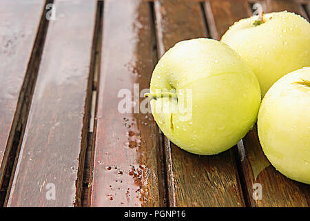 Three wet golden green apples covered by water drops on top of brown wet natural old wooden table surface. Background with ripe fruits and copy space. Stock Photo