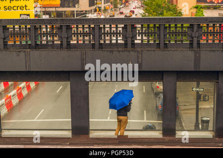 Man with a blue umbrella stands at a window on a bridge overlooking a busy NYC road. A section of The High Line walk in New York City. Stock Photo