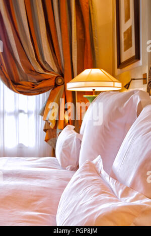 Luxury bedroom detail showing a king bed with white linens, plump pillows, and silk drapes in a beautiful light-filled room. Romantic and upscale. Stock Photo