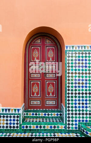 Moroccan arched exterior door, painted red with floral designs, inset in an orange plaster wall decorated with mosaic tile. Marrakech, Morocco Stock Photo