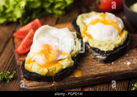 Toast with mashed avocado and poached egg on wooden serving board. Coal bread toast. Selective focus Stock Photo
