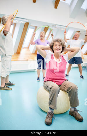 Happy senior woman is sitting on a fitness ball and doing a physical therapy exercise Stock Photo