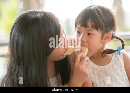 Adorable Asian child eating and sharing butter toast with mom at cafe. Outdoor family lifestyle with natural light. Stock Photo