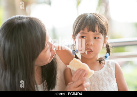 Asian child eating and sharing butter toast with mom at cafe. Outdoor family lifestyle with natural light. Stock Photo