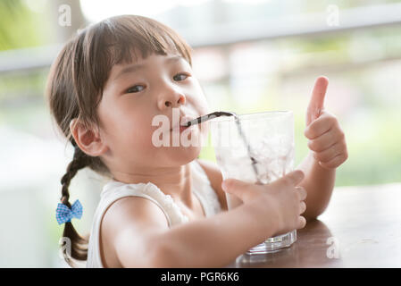 Little Asian girl drinking ice chocolate and giving thumb up at cafe. Natural light outdoor lifestyle. Stock Photo