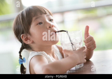 Little Asian child drinking ice chocolate and giving thumb up at cafe. Natural light outdoor lifestyle. Stock Photo