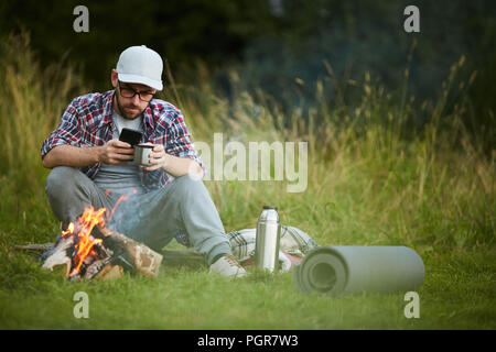 Young man drinking tea and using his smartphone by campfire Stock Photo