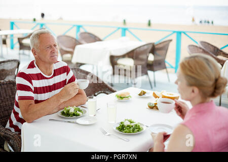 Modern senior couple having vegetarian lunch and drinking tea while sitting by table outdoors Stock Photo