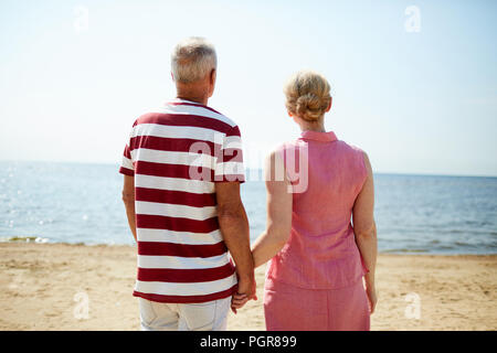 Rear view of aged couple looking at seaside while standing on sandy beach and holding by hands Stock Photo