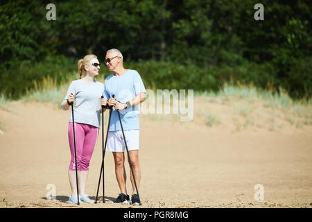 Cheerful senior active couple standing on sand and having talk while trekking on summer day Stock Photo